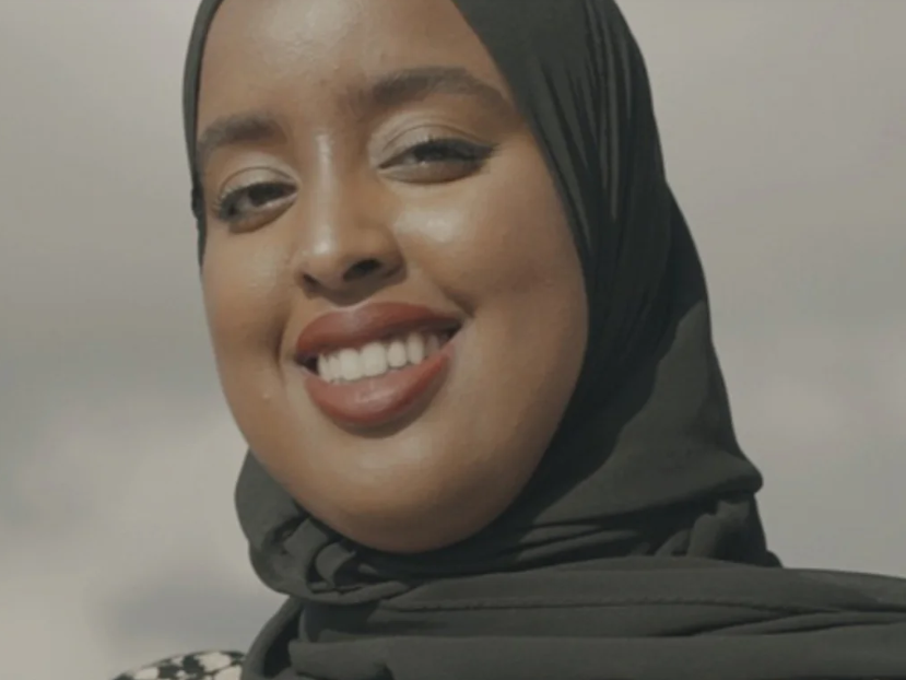 A woman wearing a black hijab smiles at the camera against a cloudy sky, reflecting the beauty of both her expression and the changing climate.