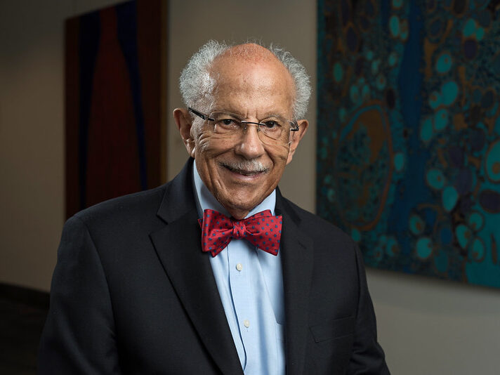An older man with grey hair, glasses, and a red bow tie smiles whilst standing indoors in front of colourful abstract artwork inspired by climate themes.