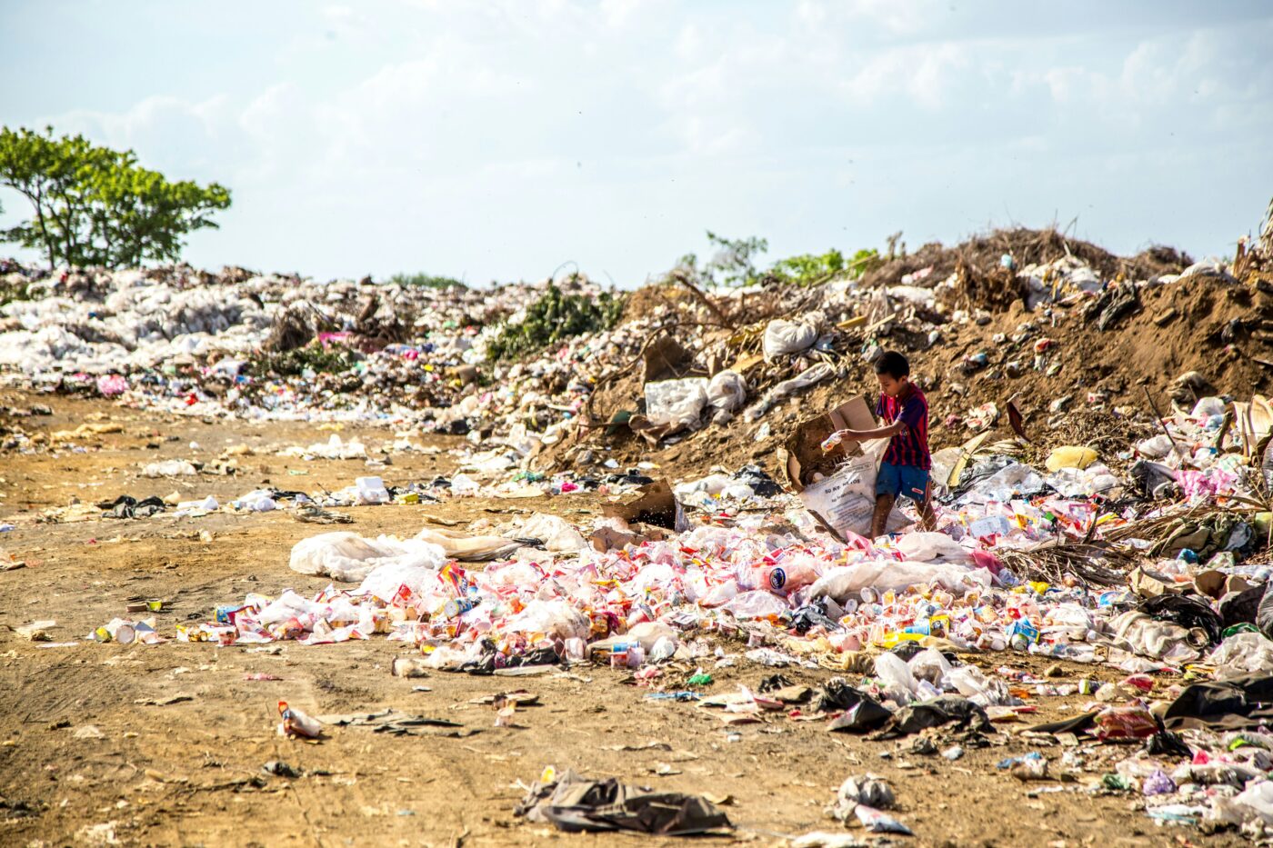 A child stands among piles of plastic waste and rubbish spread across a large open landfill under a partly cloudy sky, highlighting the environmental impact even after events like Black Friday 2024.