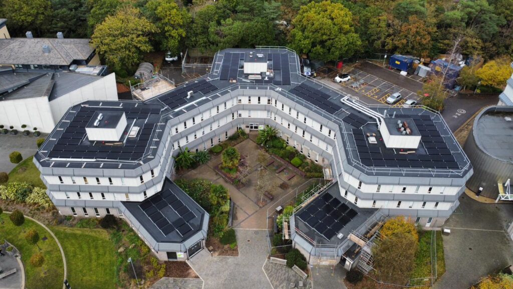 Aerial view of a modern, three-winged building with a central courtyard, surrounded by trees, greenery, and a car park.