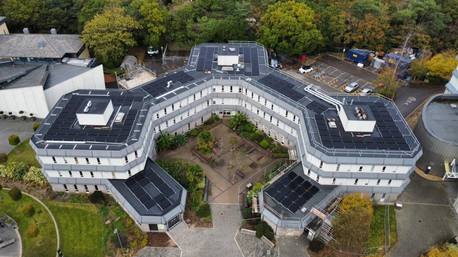 Aerial view of a modern, three-winged building with a central courtyard, surrounded by trees, greenery, and a car park.
