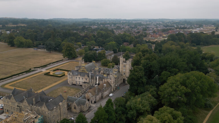 Aerial view of a large historic building surrounded by trees, with a formal garden and a residential area in the background.