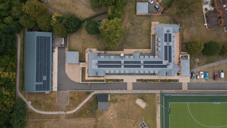 Aerial view of a school building with solar panels on the roof, adjacent sports court, and parked cars on the right-hand side.