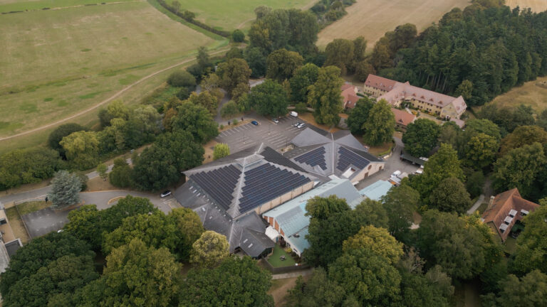 Aerial view of a large building with solar panels on the roof, surrounded by trees, car parks, and nearby residential buildings in a rural area.