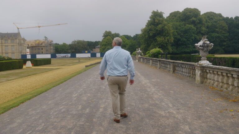 A man in a light blue shirt and beige trousers walks alone on a wide gravel path beside a stone balustrade, with trees and a construction crane in the background.
