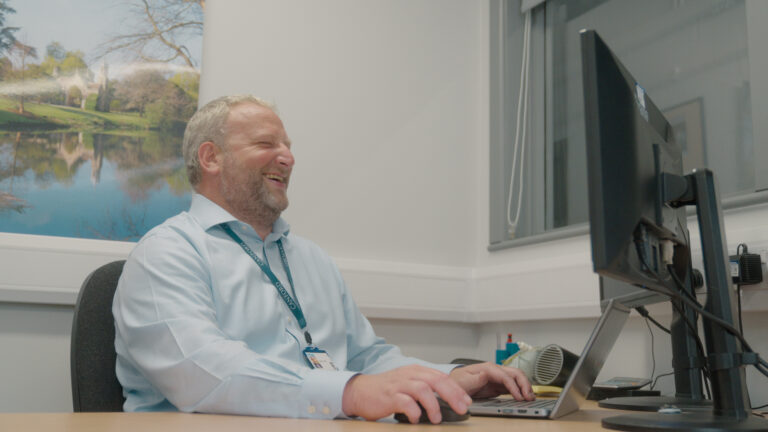 A man with a lanyard sits at a desk, smiling whilst working on a computer in an office with a landscape photo on the wall.