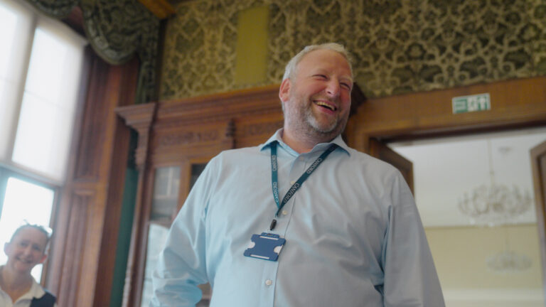 A man wearing a blue shirt and a lanyard smiles whilst standing indoors near an ornate wooden cupboard, with another person blurred in the background.