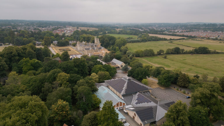 Aerial view of a large historic building surrounded by trees and gardens, with open fields and a car park in the foreground, under an overcast sky.