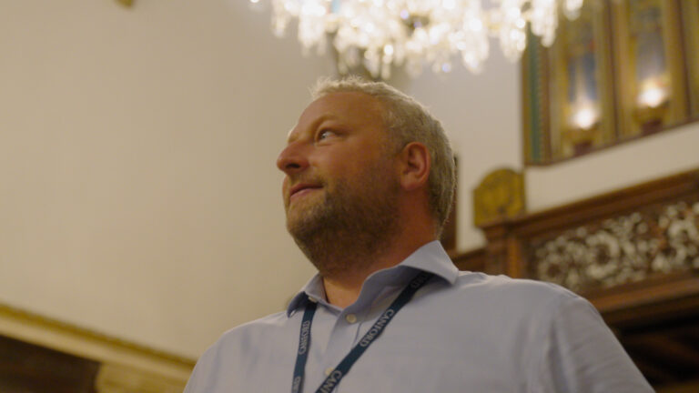 A man wearing a light blue shirt and a lanyard stands indoors under a chandelier, with ornate woodwork visible in the background.