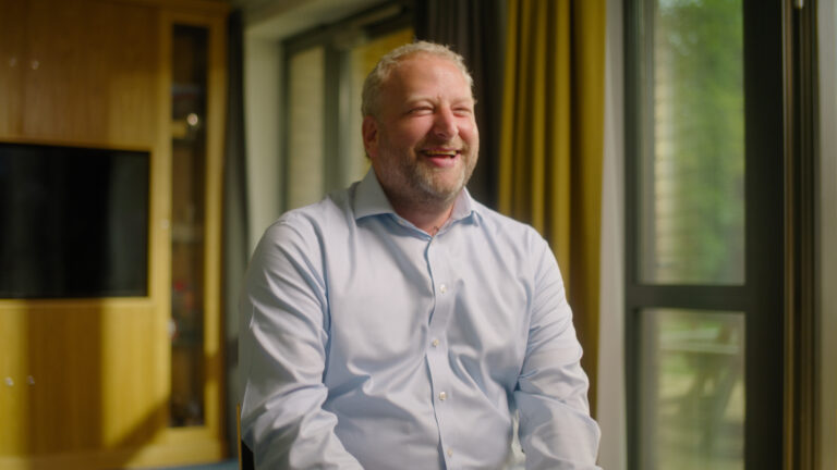A man with fair hair and a beard, wearing a light blue shirt, sits indoors on a chair, smiling. Large windows with trees outside are in the background.