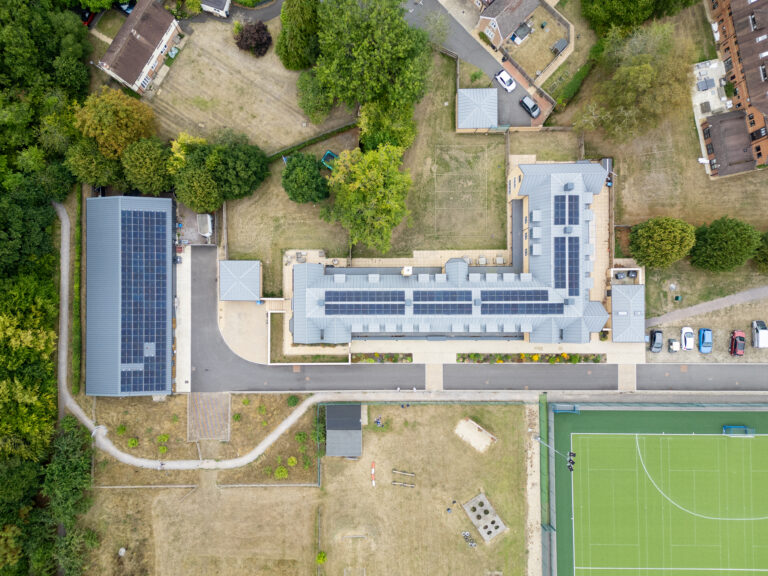Aerial view of a building complex with solar panels on the roofs, surrounded by trees, parking areas, and a sports pitch.