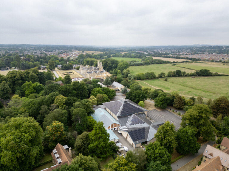 Aerial view of a rural area with a large building complex surrounded by trees, open fields, and a church visible in the background under a cloudy sky.