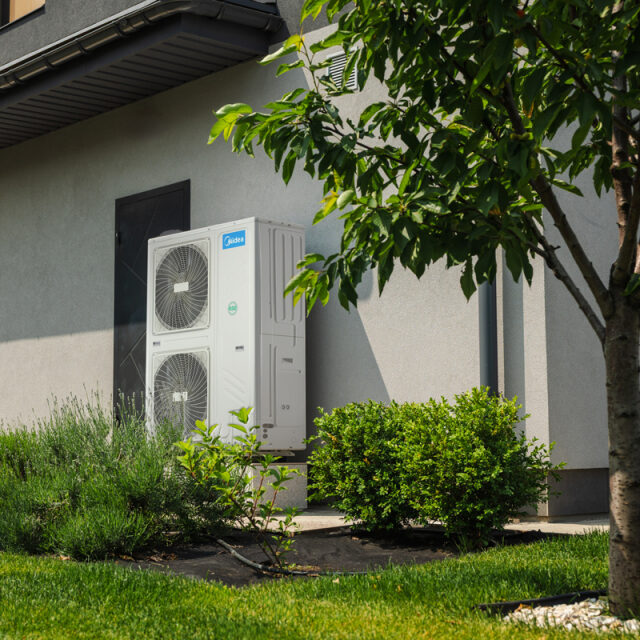 Two outdoor heat pump units are installed against the exterior wall of a modern house, surrounded by green shrubs and grass.