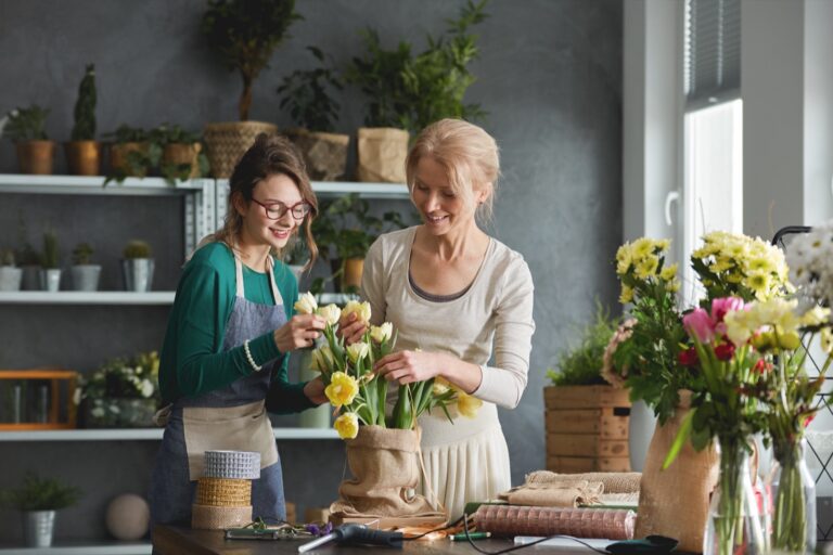 Two women arrange flowers together at a table in a room filled with plants and floral supplies.