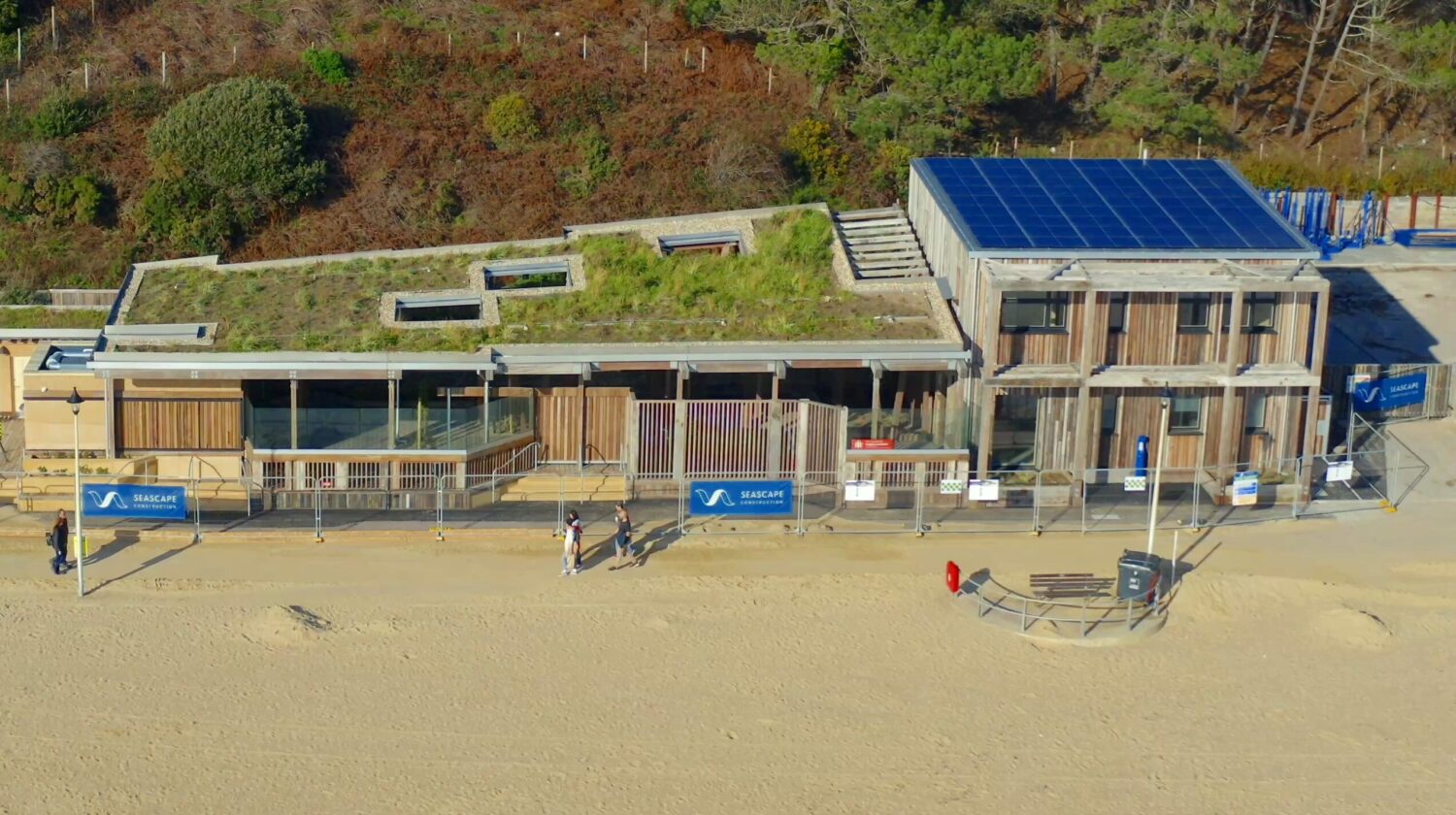 A modern, eco-friendly building with a green roof and solar panels sits beside a sandy beach, bordered by a fence, with several people walking nearby.