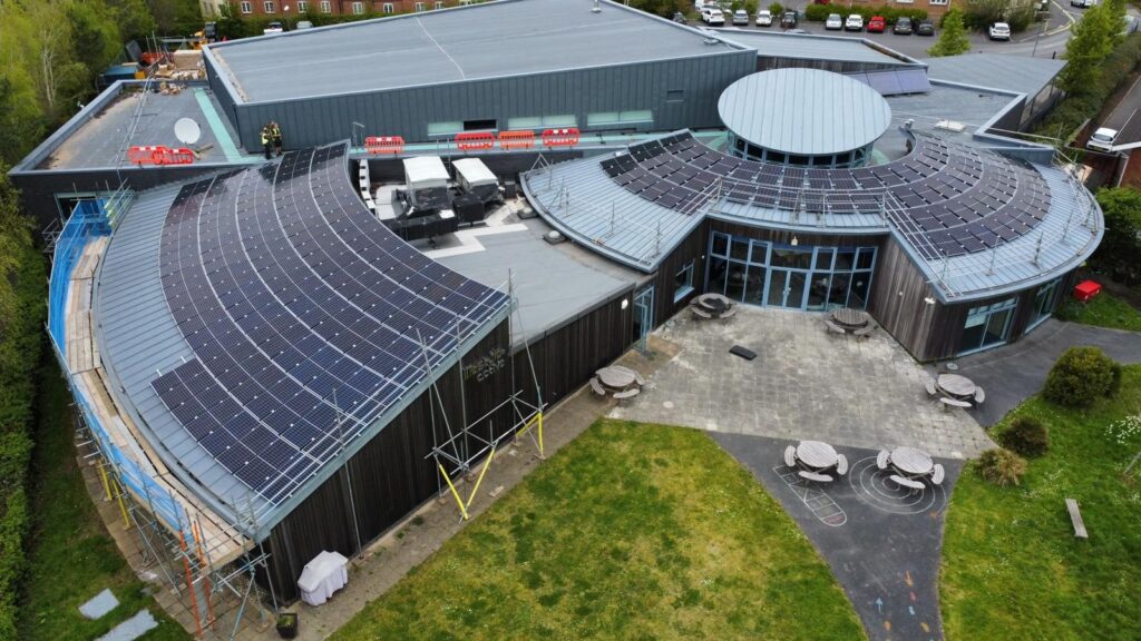 Aerial view of a modern building with extensive solar panels on the roof, surrounded by grass, paved footpaths, and outdoor seating areas.