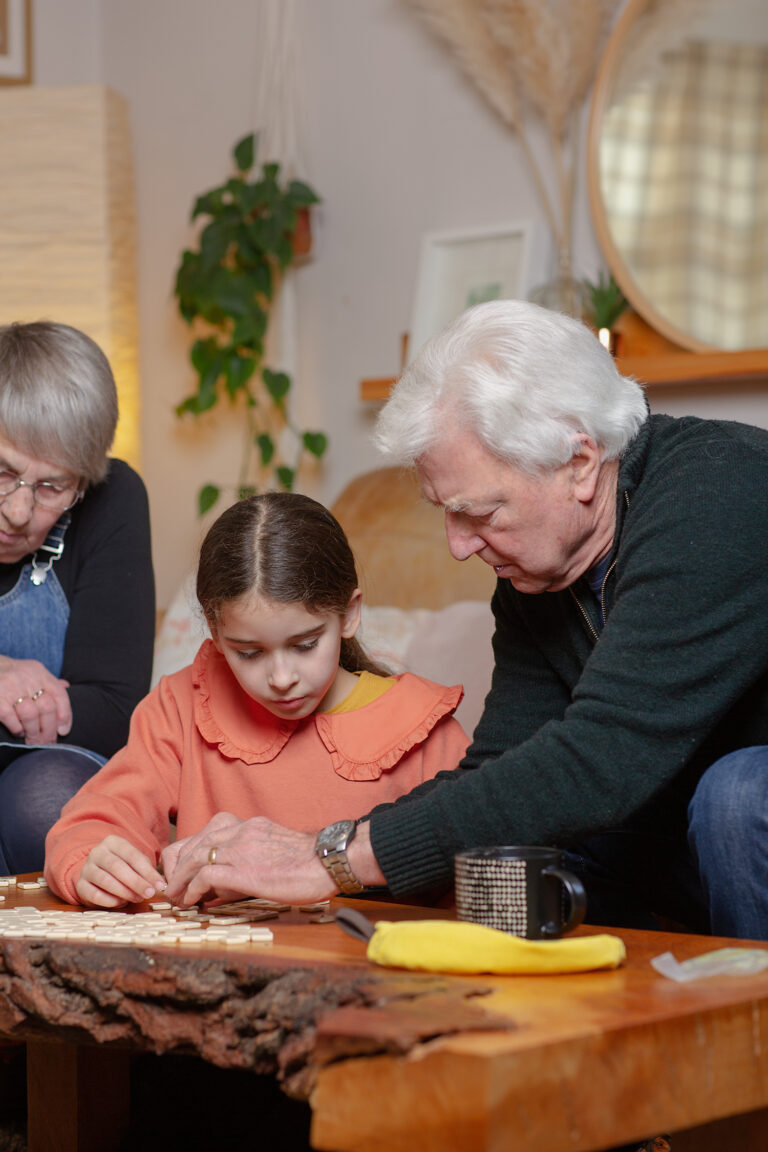 An older man and a young girl sit at a wooden table, focussing on a board game; an older woman watches them. A coffee mug and yellow cloth are on the table.