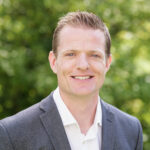 A man in a grey suit jacket and white dress shirt smiles at the camera with greenery in the background.