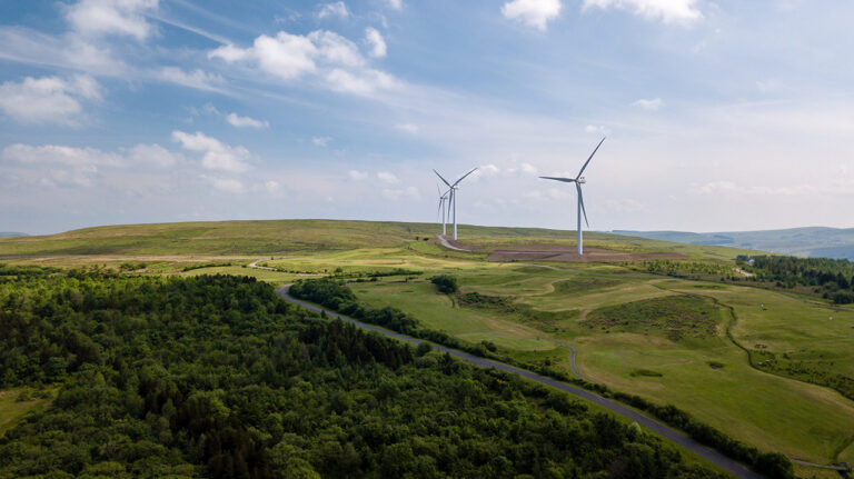 A landscape with rolling green hills and a forested area features two large wind turbines generating renewable electricity under a partly cloudy sky.