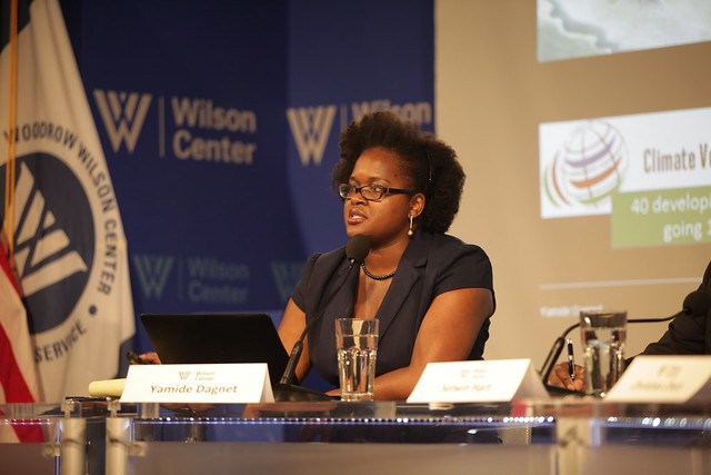 A woman, identified as Yamide Dagnet, speaks into a microphone at a panel discussion for International Women’s Day at the Wilson Centre. A presentation and nameplates are visible in the background.