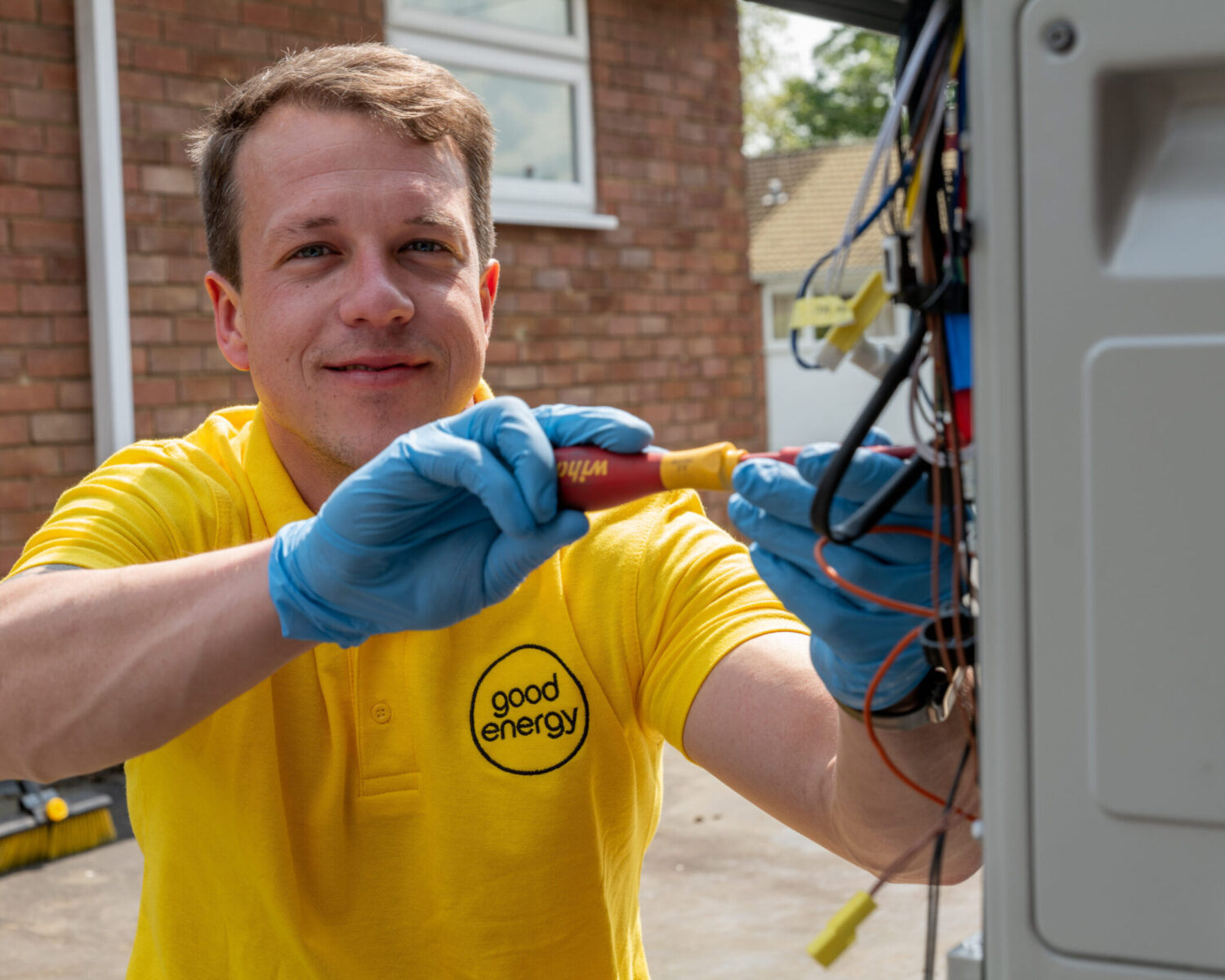A heat pump technician in a yellow good energy shirt and blue gloves works on electrical wiring outdoors, ready to share home heating tips to improve comfort and efficiency.