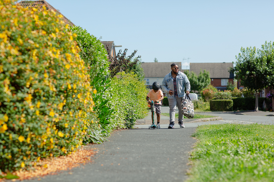 An adult walks beside a child riding a scooter on a suburban pavement lined with greenery and yellow flowers, embracing eco-friendly living with eco-friendly transport on a sunny day.