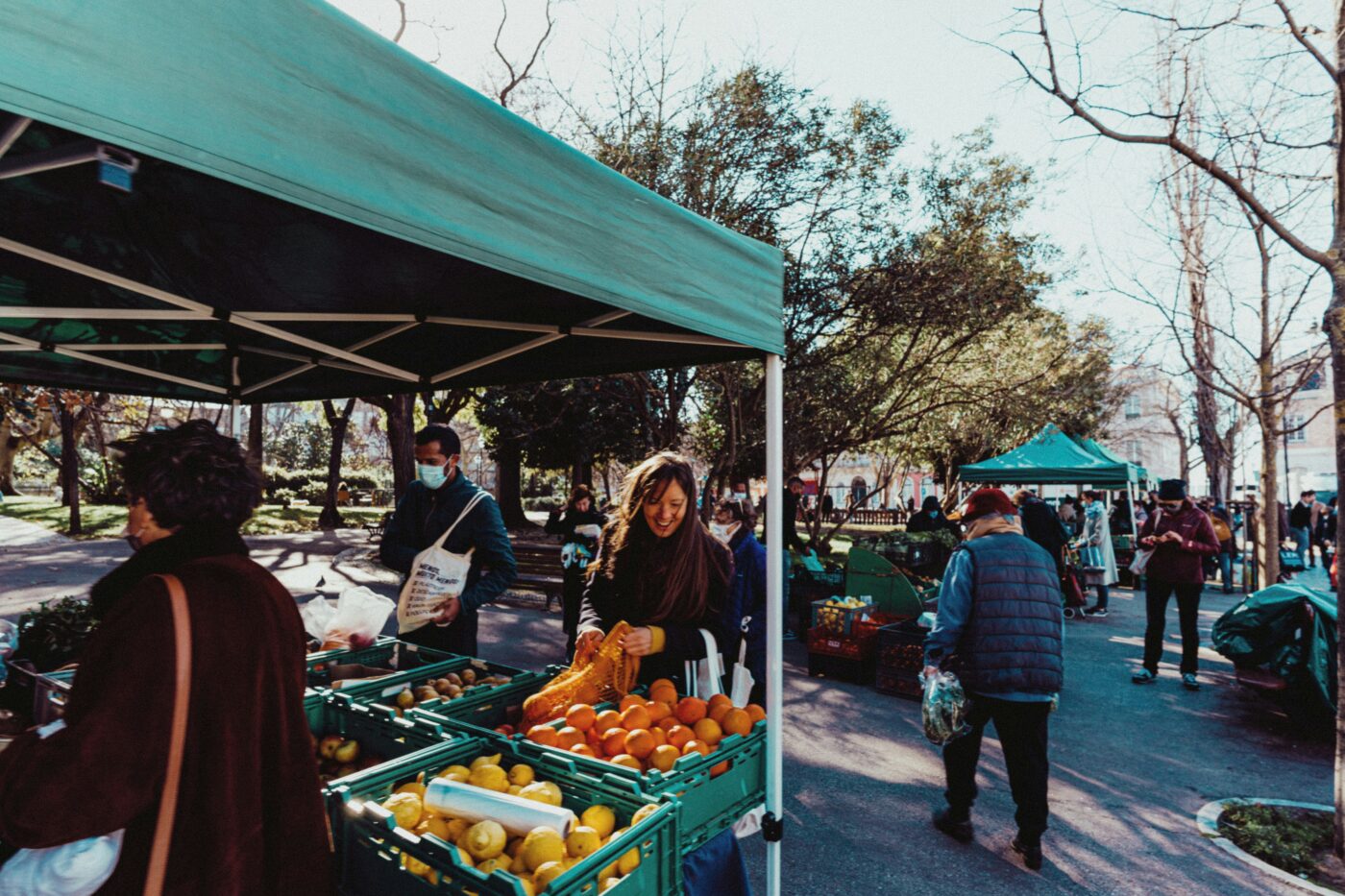 People shop for fresh produce at outdoor market stalls under green canopies on a sunny day; fruit and vegetables are displayed in green crates, reflecting a commitment to buying local and eco-friendly living.