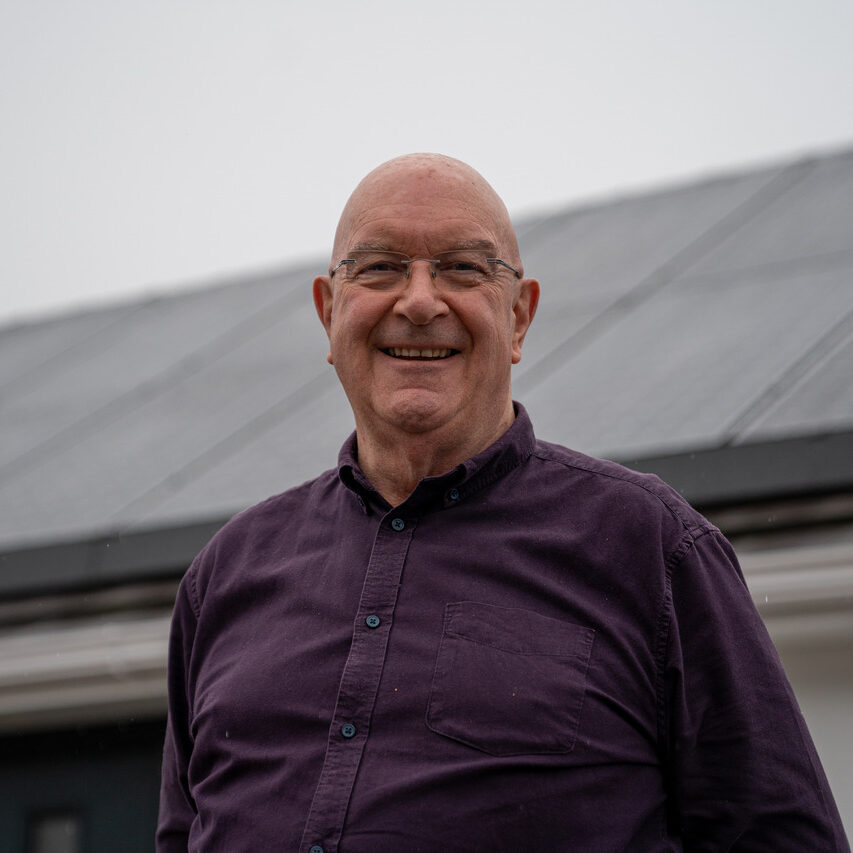 A man wearing glasses and a dark purple shirt stands outside, smiling, with a building, cloudy sky, and signs of recent solar panel installation in the background.