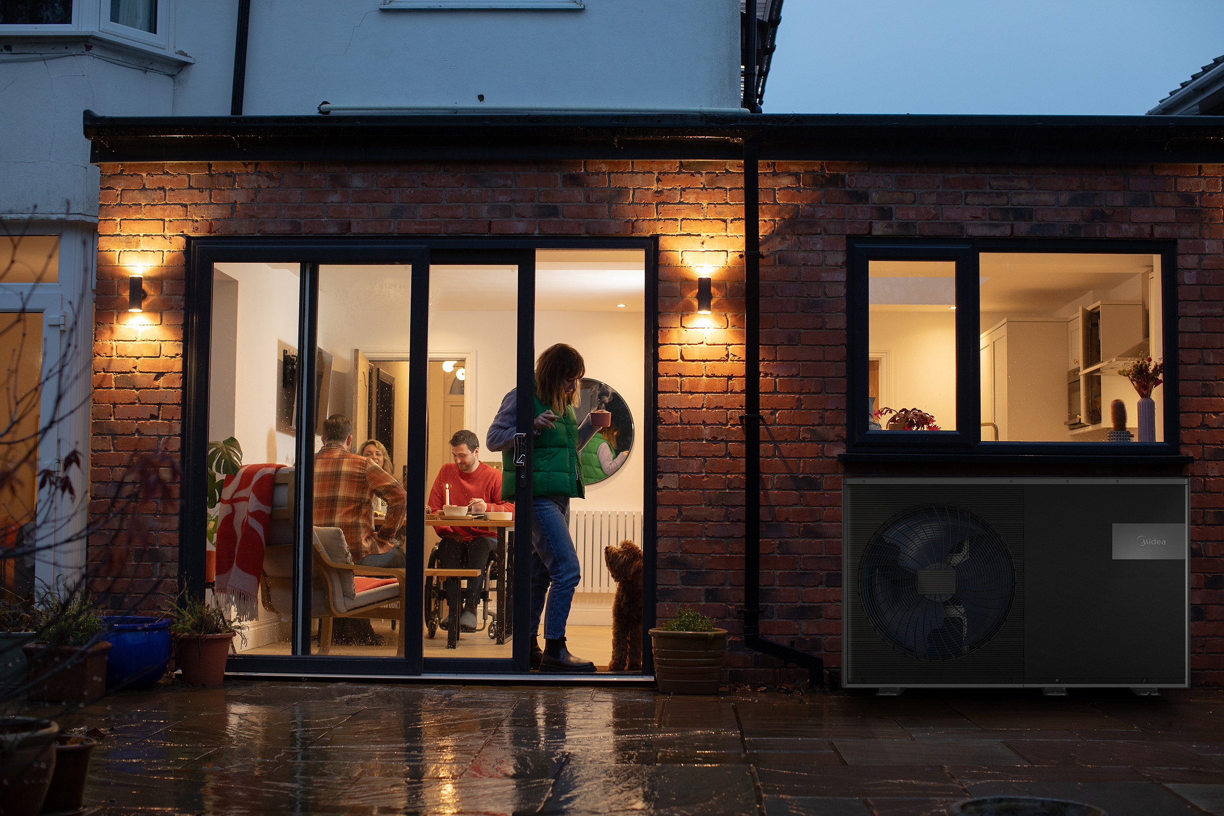 A woman stands by a glass door with a dog, whilst three people sit at a dining table inside a brick house on a rainy evening.