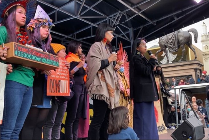 A group of women stand on stage holding placards and a microphone, addressing a crowd about climate action at an outdoor event near a statue and historic buildings.
