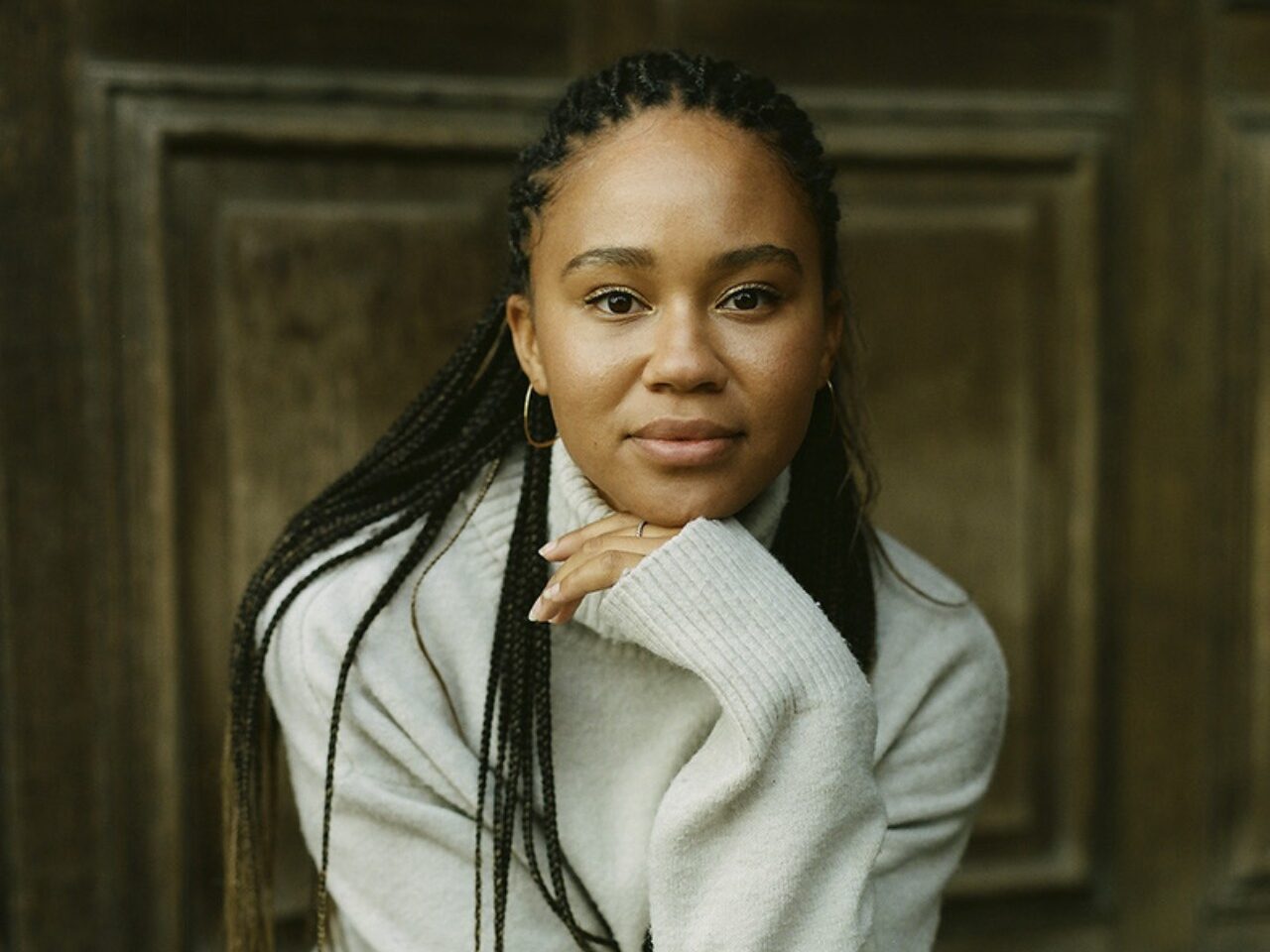 A person with long plaits, wearing a light jumper and jeans suitable for the mild climate, sits on a bench in front of a wooden door, resting their chin on their hand and looking at the camera.