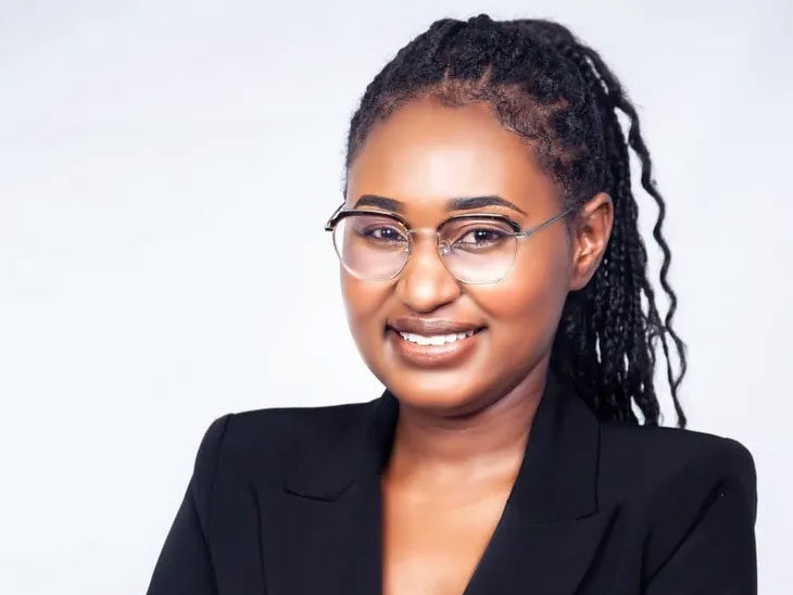Woman with glasses and plaited hair, wearing a black blazer, smiles with arms crossed against a plain light background, radiating confidence and a passion for climate advocacy.