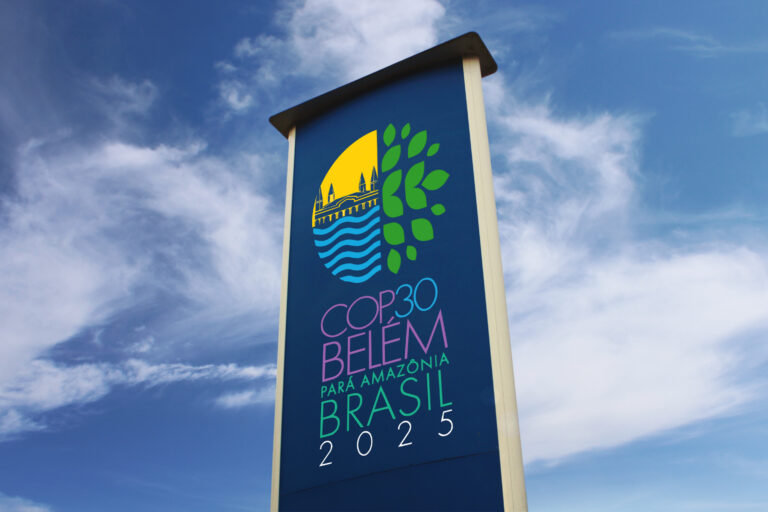 A tall sign displays the logo and text for COP30 Belém for the Amazon, Brazil 2025, set against a blue sky with clouds, highlighting the global journey from COP29 to this landmark event.