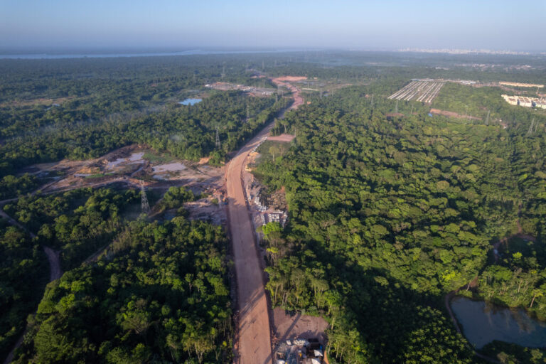 Aerial view of a road under construction cutting through a dense forest, with cleared land and construction equipment visible along the route.