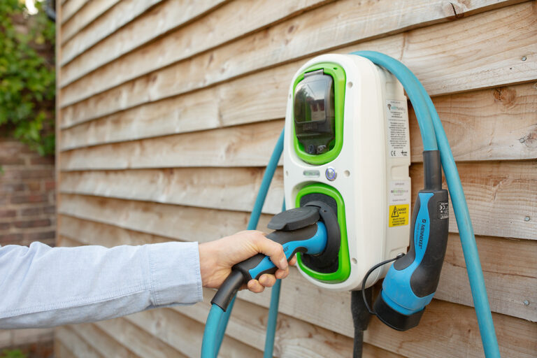 A person plugs a blue charging cable into an electric vehicle charging station mounted on a wooden wall.