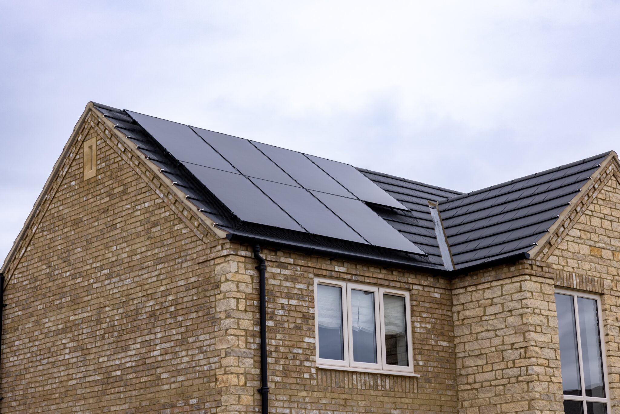 A tan brick house with dark grey roof tiles, featuring several black solar panels fitted on the roof above two windows.