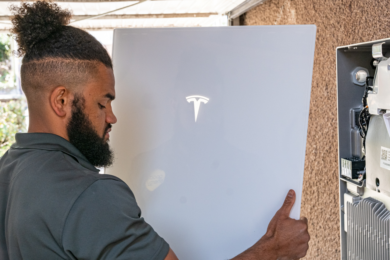 A person installs a Tesla Powerwall battery unit on an exterior wall next to an open control panel.