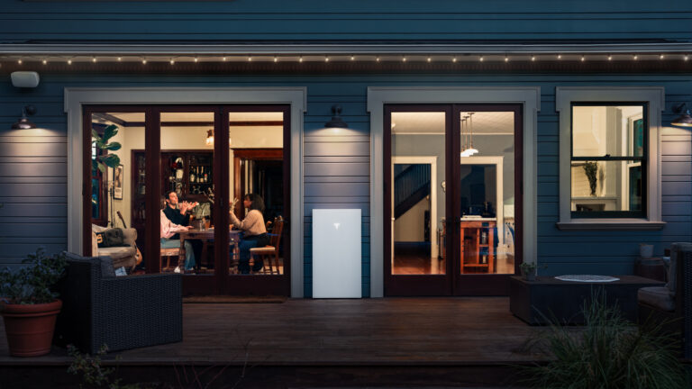 A Tesla Powerwall is mounted on the exterior wall of a house with large windows showing people sitting and eating inside.