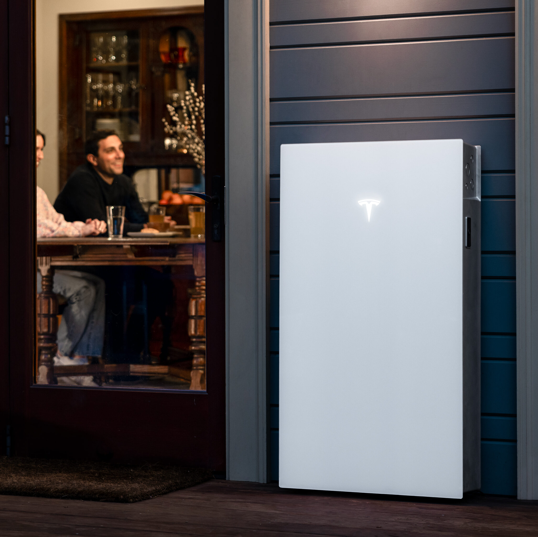 A Tesla Powerwall battery is mounted on an exterior wall next to an open glass door, with two people visible inside a cosy, well-lit room.