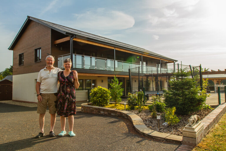 An older man and woman stand smiling in front of a modern two-storey house with a balcony and landscaped garden on a sunny day.